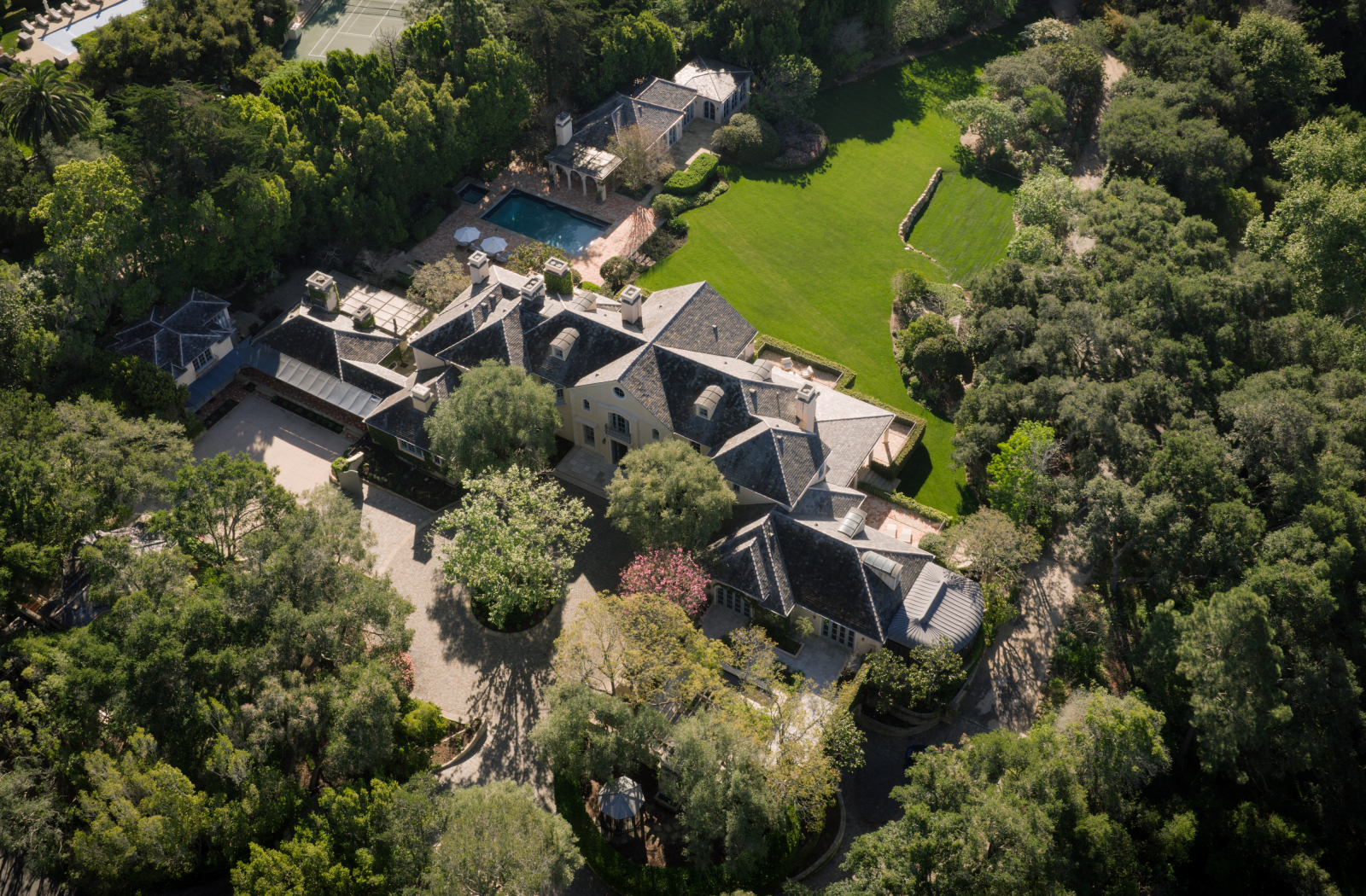 Aerial view of a large luxury estate surrounded by trees, featuring a multi-wing home, expansive lawn, swimming pool, and landscaped grounds.